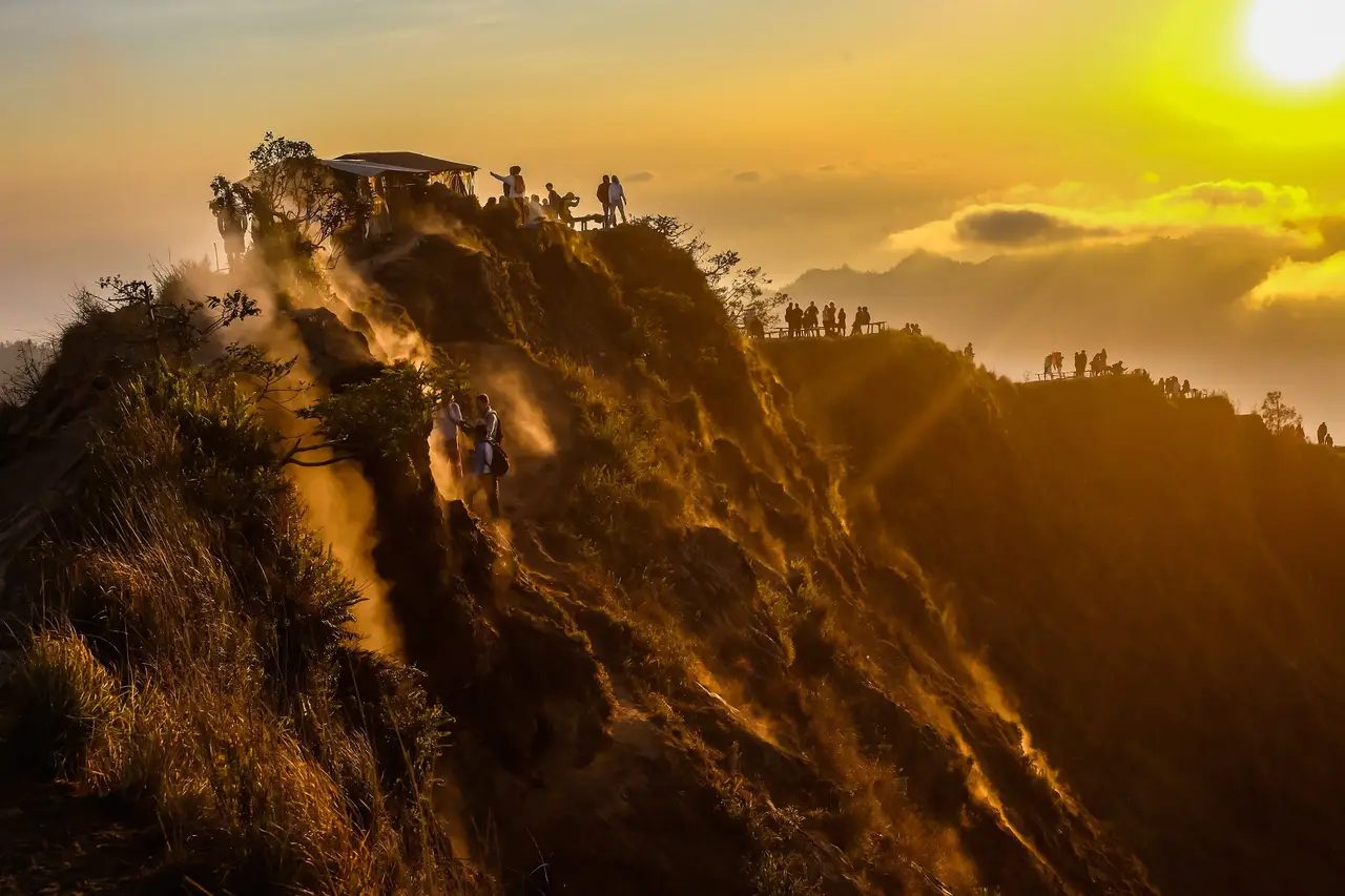 The ridge of Mount Batur lit gold at sunrise, with a line of tiny hikers silhouetted on the summit viewpoint.