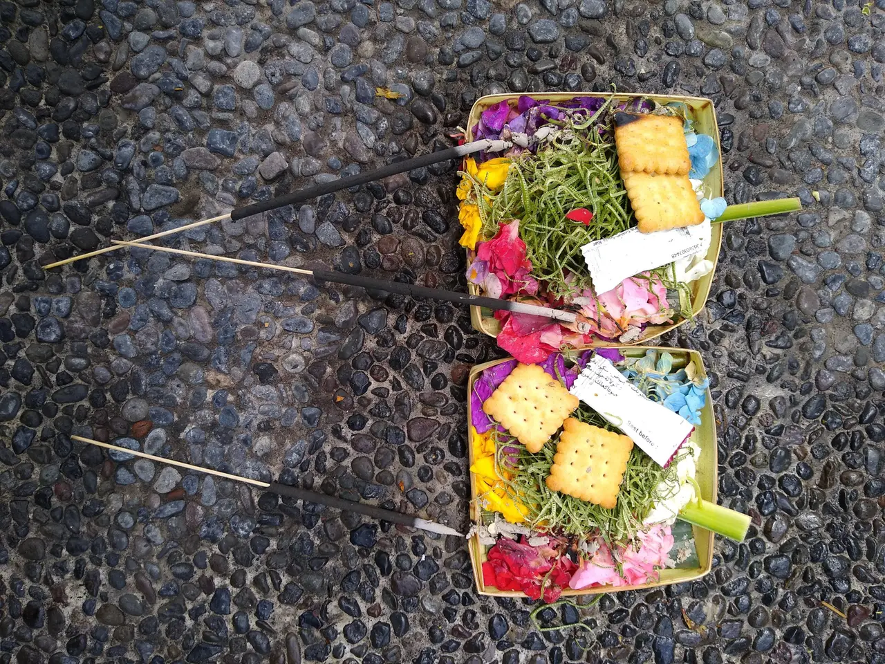 Two canang sari offerings of flowers and a biscuit in small palm-leaf trays on dark pebbles, with incense sticks laid beside them.