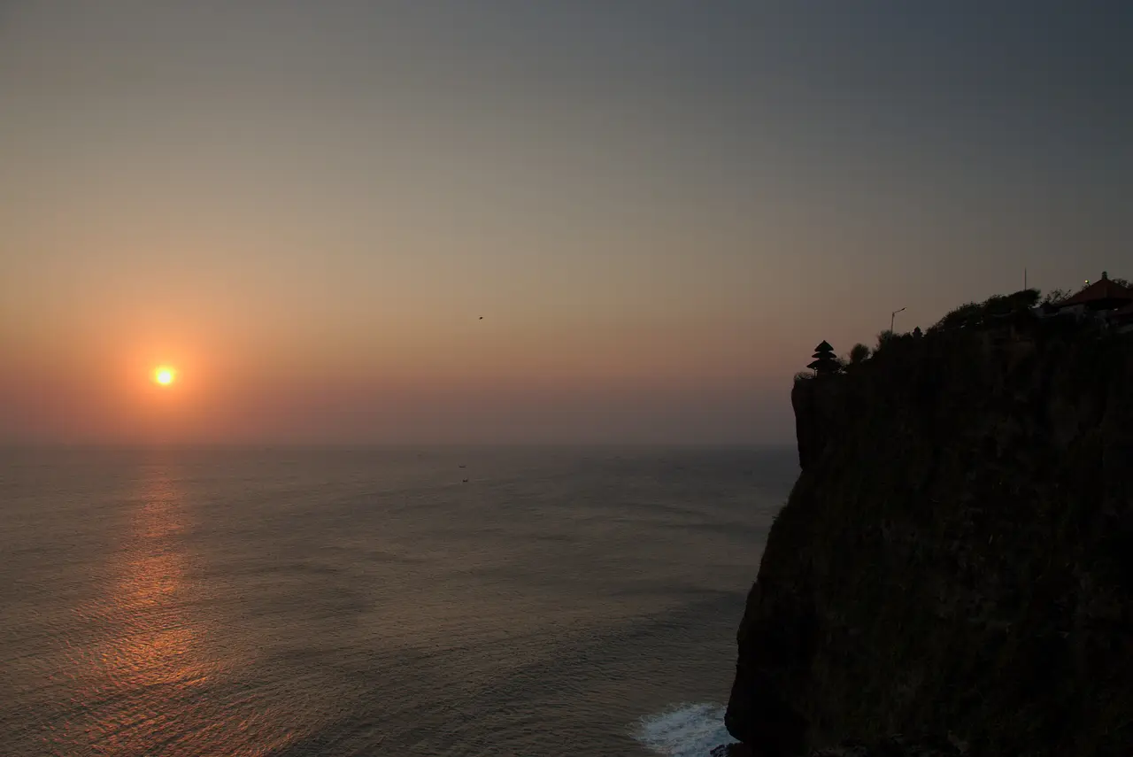 The sun setting over the Indian Ocean with a path of orange light on the water, and Uluwatu Temple on its sheer cliff at the right edge.